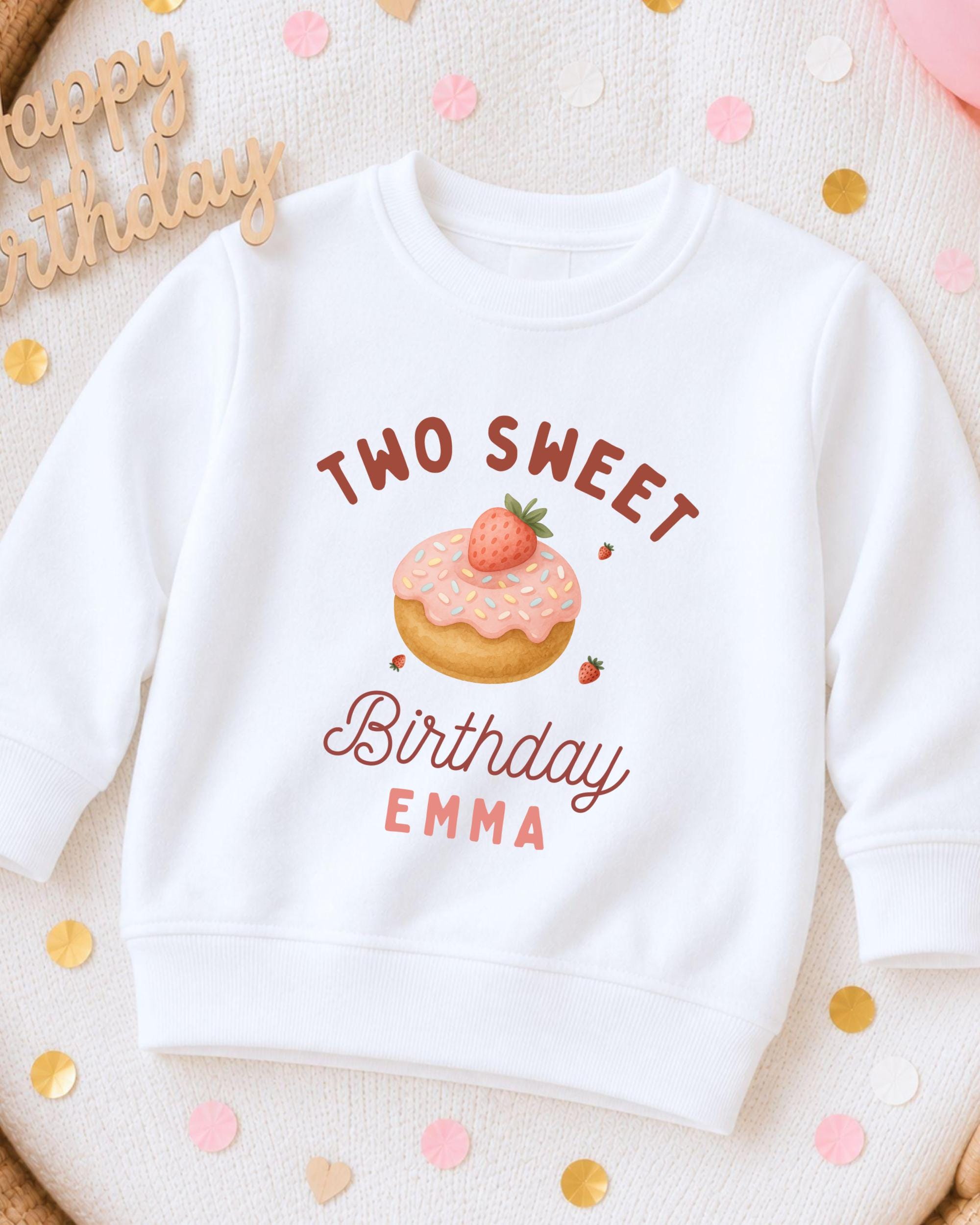 A white sweatshirt with a birthday-themed design, including a donut with a strawberry and the text "Two Sweet Birthday Emma".