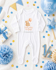A white onesie with a teddy bear and the text "Half Way to One" is displayed among various baby-themed items, including a blue balloon, confetti, and a star decoration.