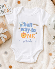 A white onesie with a cartoon character and text printed on it, placed on a white blanket with confetti around it.