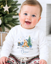 A smiling baby boy wearing a white sweater with a Christmas-themed design and text, sitting in front of a Christmas tree.