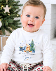 A smiling baby boy wearing a white sweater with a Christmas-themed design and text, sitting in front of a Christmas tree.