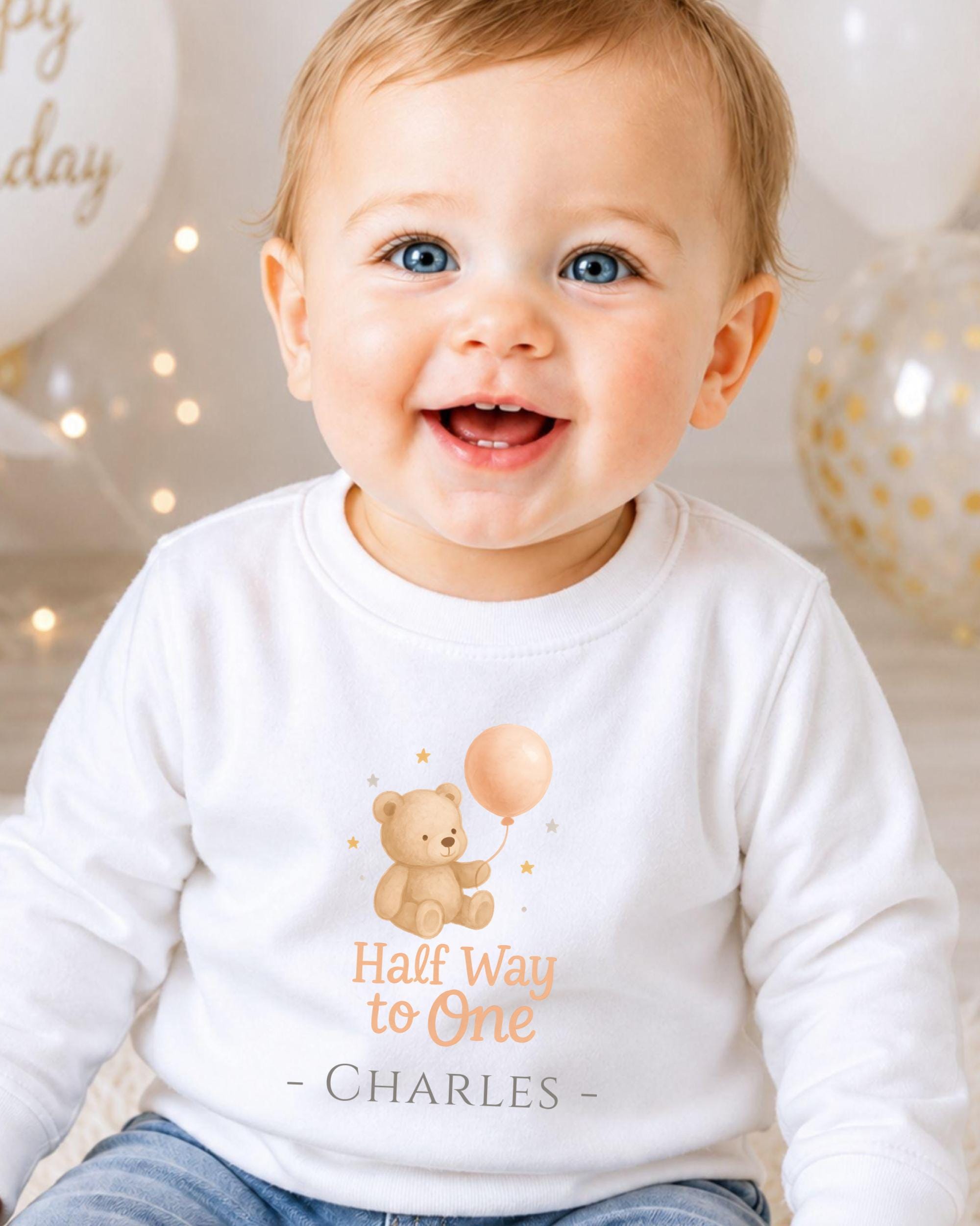 A smiling baby boy wearing a white shirt with a teddy bear and a balloon, sitting in front of birthday balloons.