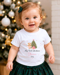 A smiling baby girl wearing a white t-shirt with a Christmas tree and presents, standing in front of a Christmas tree with ornaments.