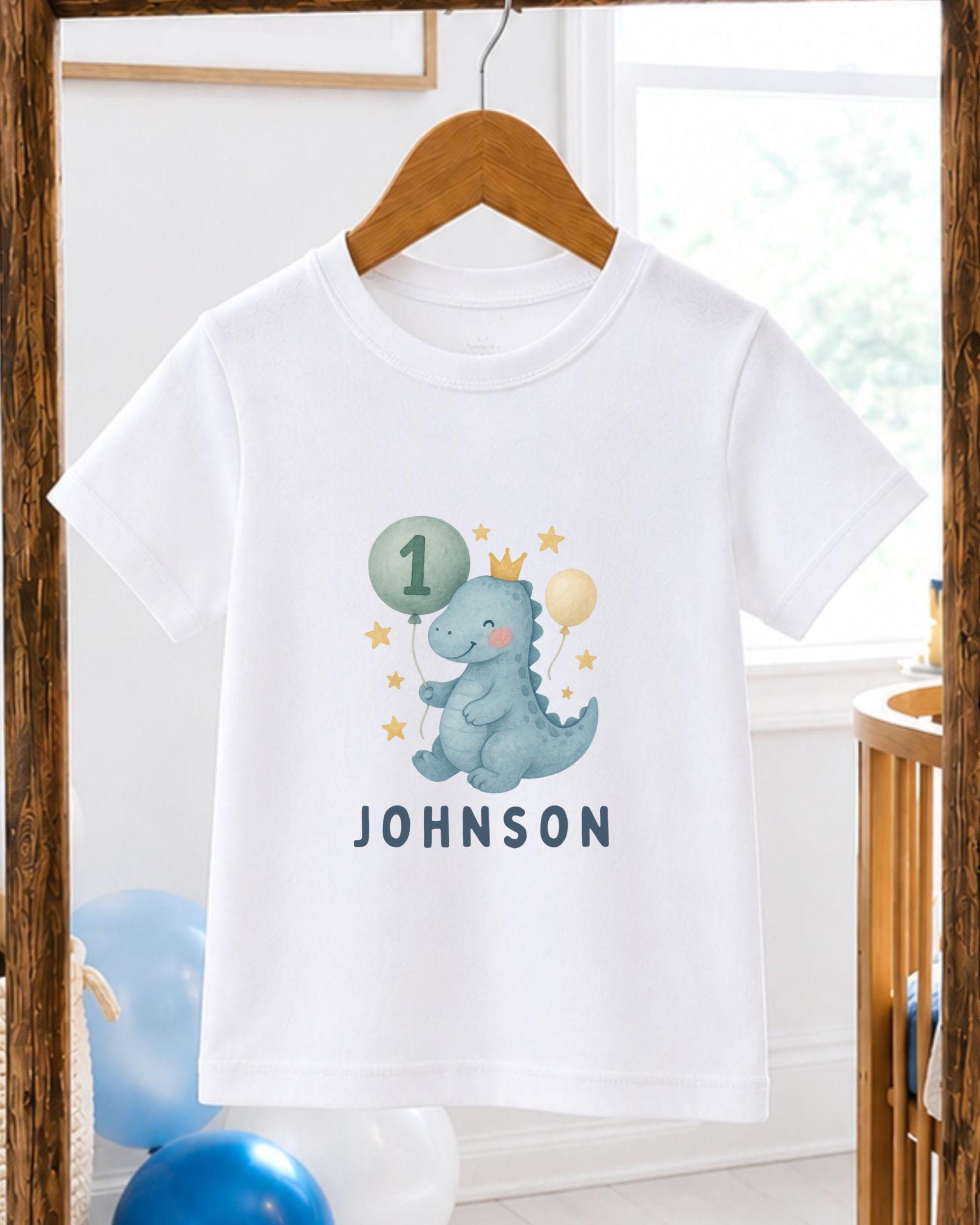 A white t-shirt with a cartoon dinosaur wearing a crown and holding balloons, along with the text "JOHNSON" printed on it.