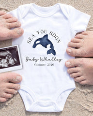 A white onesie with a graphic of a whale and the text "SEA YOU SOON Baby Whalley Summer 2026" is displayed on a sandy beach, surrounded by people's hands.