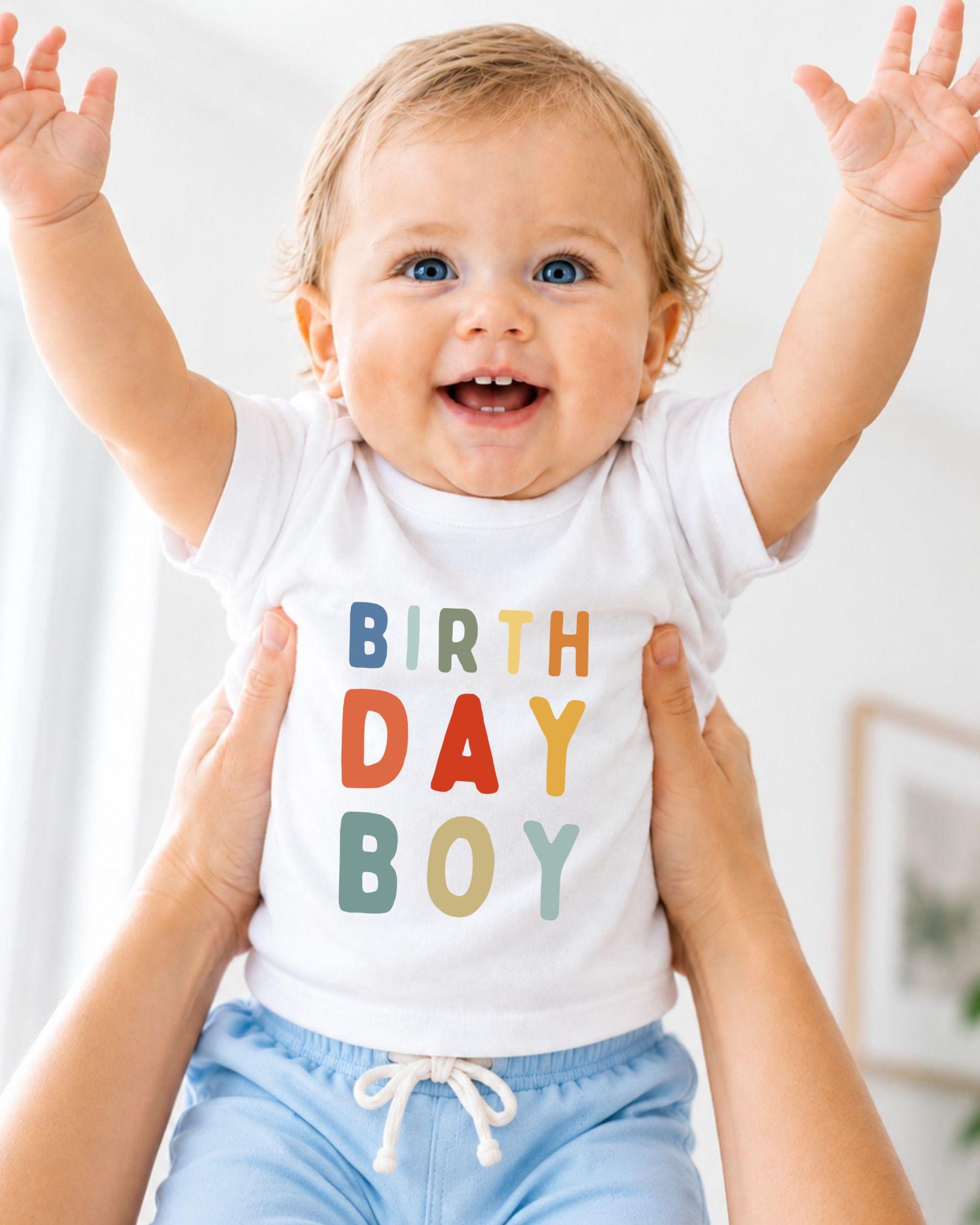 A smiling baby wearing a white t-shirt with the text "BIRTHDAY BOY" is being held up by someone's arms.