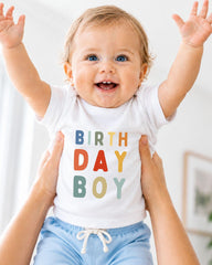 A smiling baby wearing a white t-shirt with the text "BIRTHDAY BOY" is being held up by someone's arms.