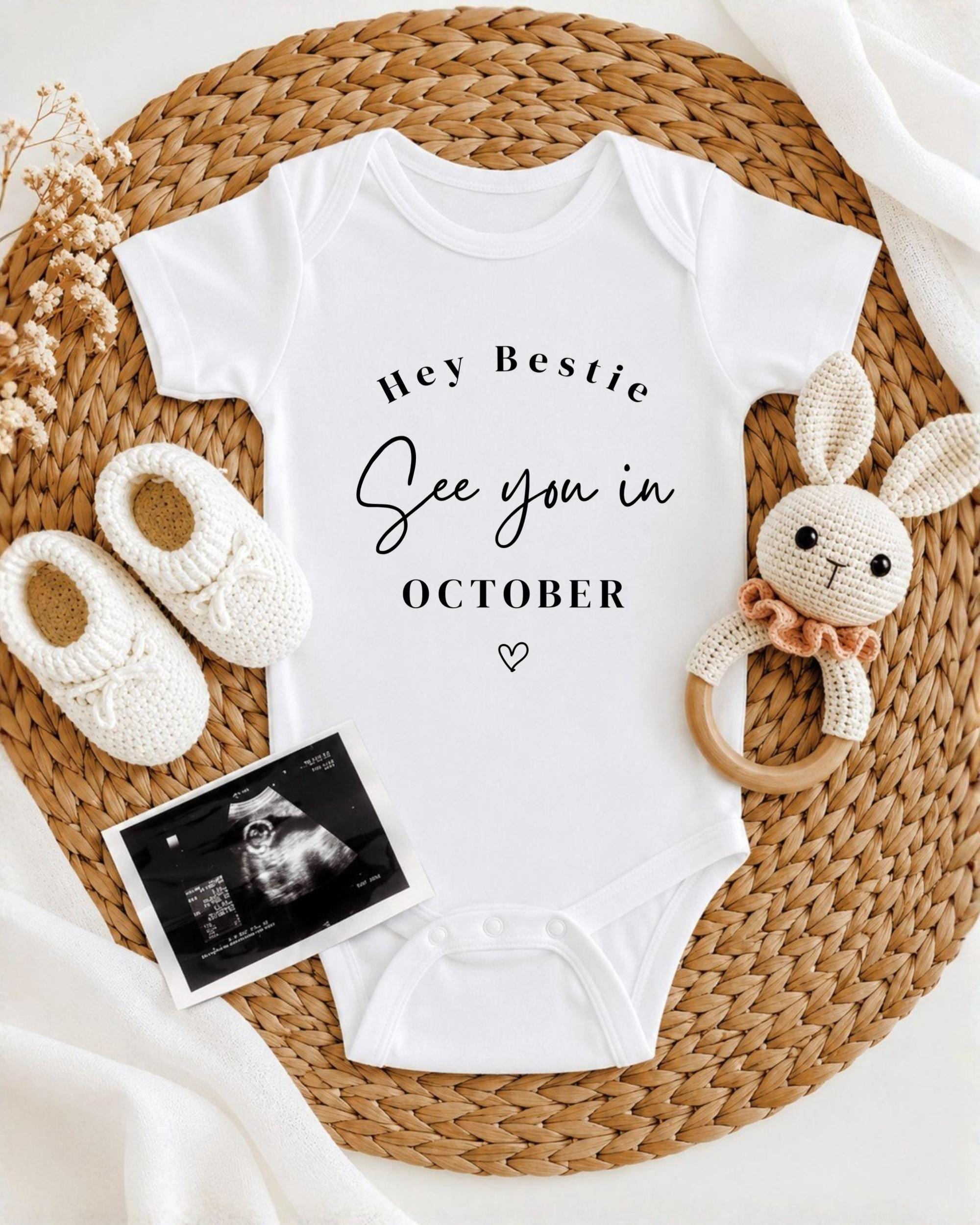A baby onesie with the text "Hey Bestie See you in October" printed on it, along with a pair of white crocheted shoes and a crocheted rabbit toy. The items are arranged on a woven placemat, creating a cozy and welcoming display.