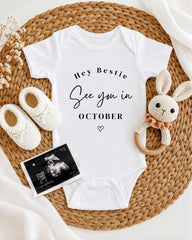 A baby onesie with the text "Hey Bestie See you in October" printed on it, along with a pair of white crocheted shoes and a crocheted rabbit toy. The items are arranged on a woven placemat, creating a cozy and welcoming display.