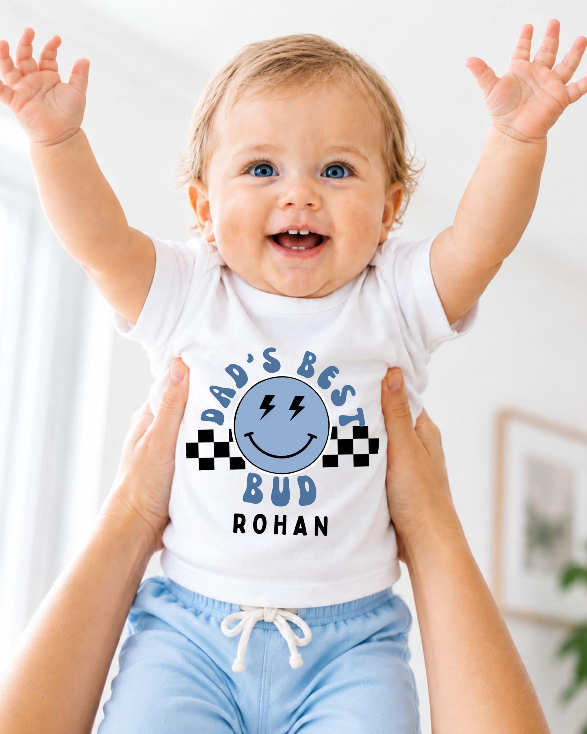 A smiling baby wearing a white shirt with a graphic and text, being held up by someone's arms.