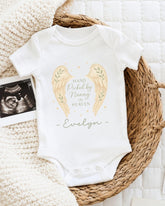 A white onesie with a design of angel wings and the text "Hand picked by Nancy in Heaven" is displayed on a bed, accompanied by a photograph of an ultrasound image.