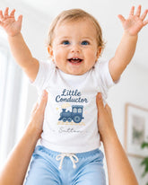 A smiling baby wearing a white shirt with a train graphic and the name "Sutton" printed on it, being held up by someone's arms.
