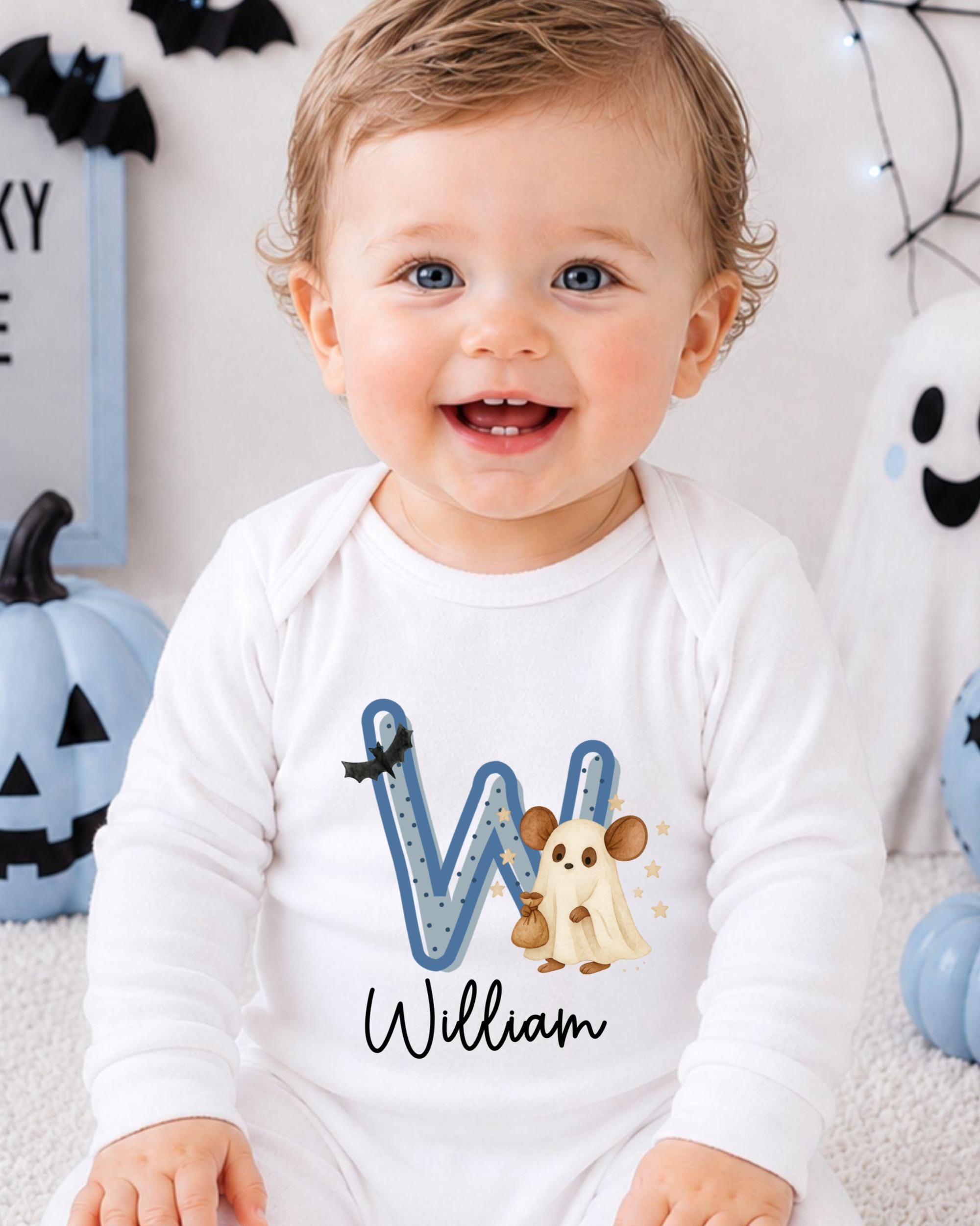 A smiling baby boy wearing a white onesie with a letter "W" and a ghost character on it, sitting in front of Halloween decorations.