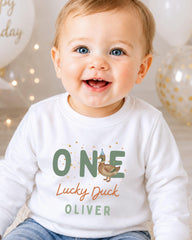 A smiling baby boy wearing a white shirt with the text "ONE Lucky Duck Oliver" printed on it.