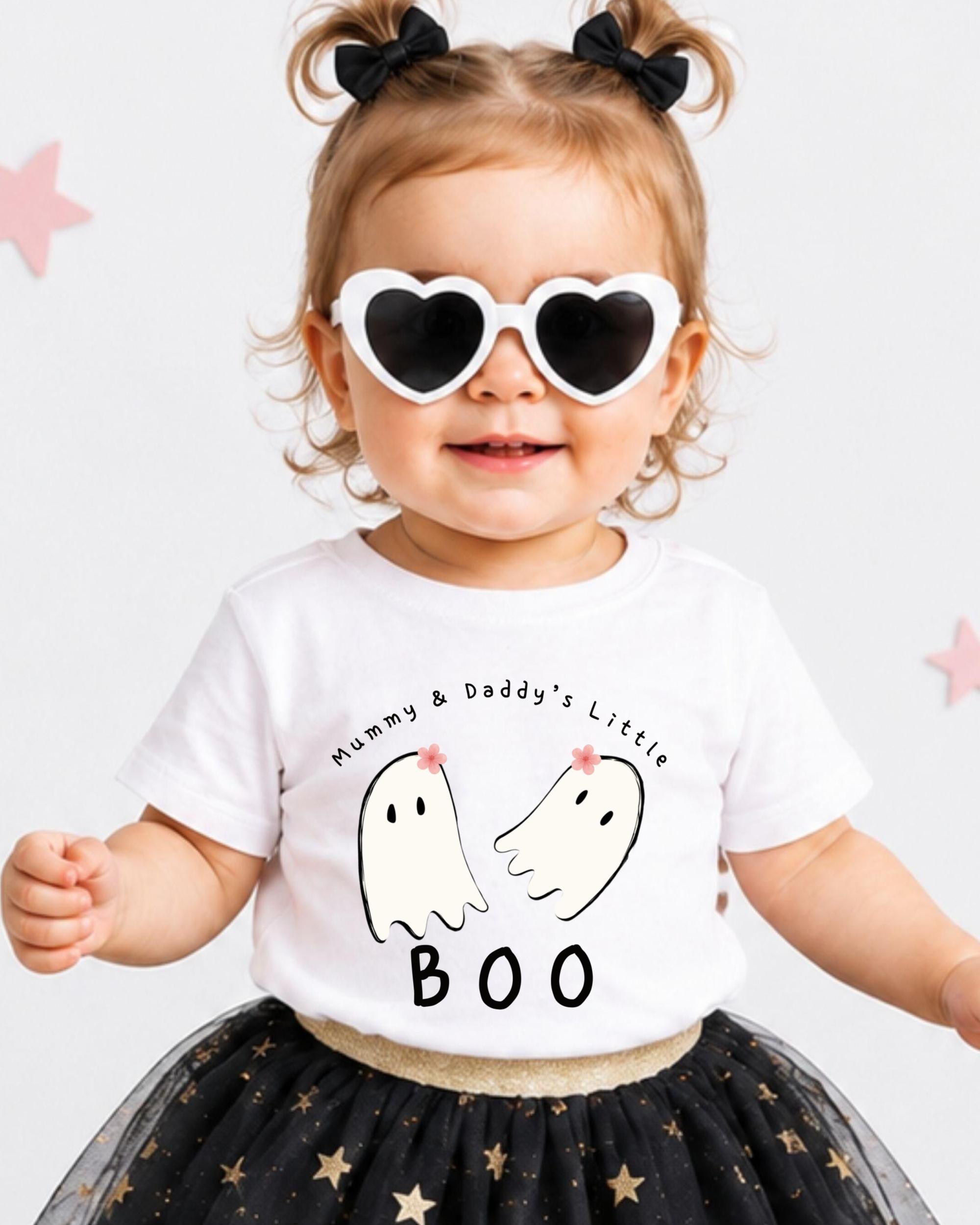 A young girl wearing a white t-shirt with a graphic of two ghostly figures and the text "Mummy & Daddy's Little" and "BOO" printed on it, along with heart-shaped sunglasses. She is also wearing a black and gold tutu skirt.