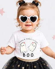 A young girl wearing a white t-shirt with a graphic of two ghostly figures and the text "Mummy & Daddy's Little" and "BOO" printed on it, along with heart-shaped sunglasses. She is also wearing a black and gold tutu skirt.
