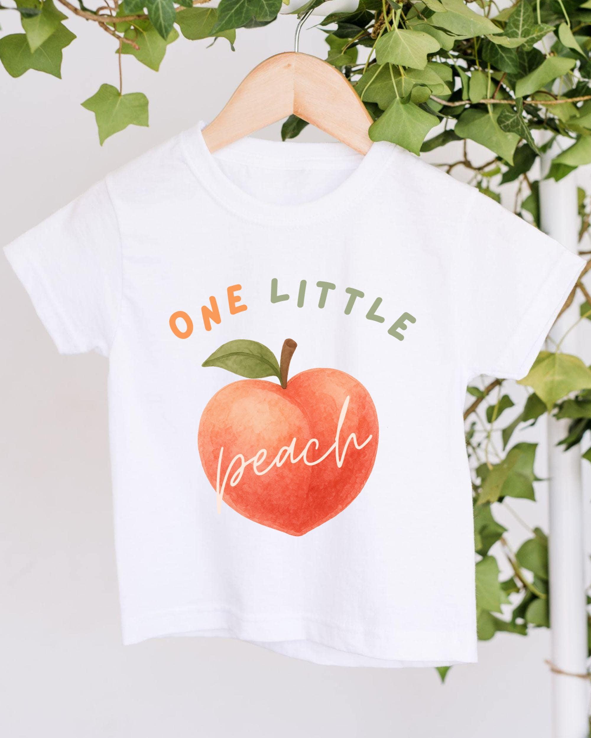 A white t-shirt with a graphic design of an orange peach and the text "ONE LITTLE peach" printed on it, hanging on a wooden hanger.