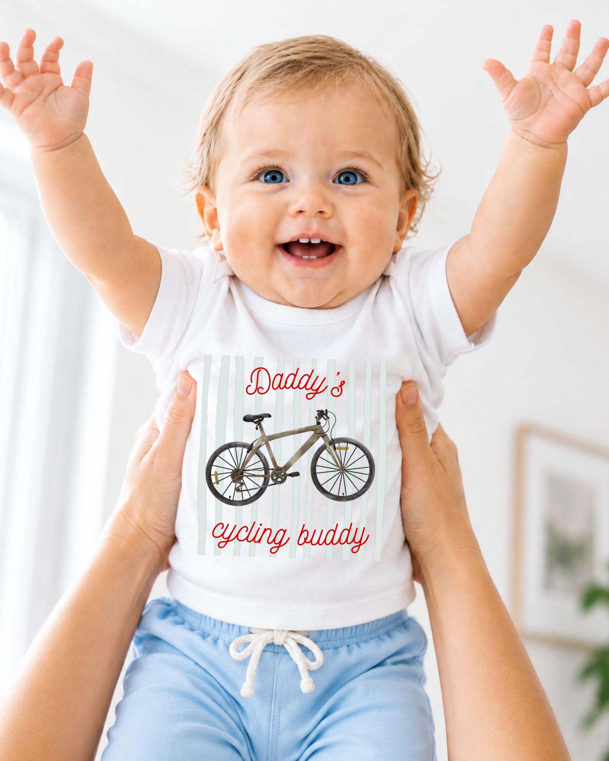 A smiling baby wearing a white shirt with a bicycle graphic and the text "Daddy's cycling buddy" being held up by someone's arms.