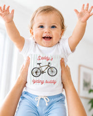 A smiling baby wearing a white shirt with a bicycle graphic and the text "Daddy's cycling buddy" being held up by someone's arms.
