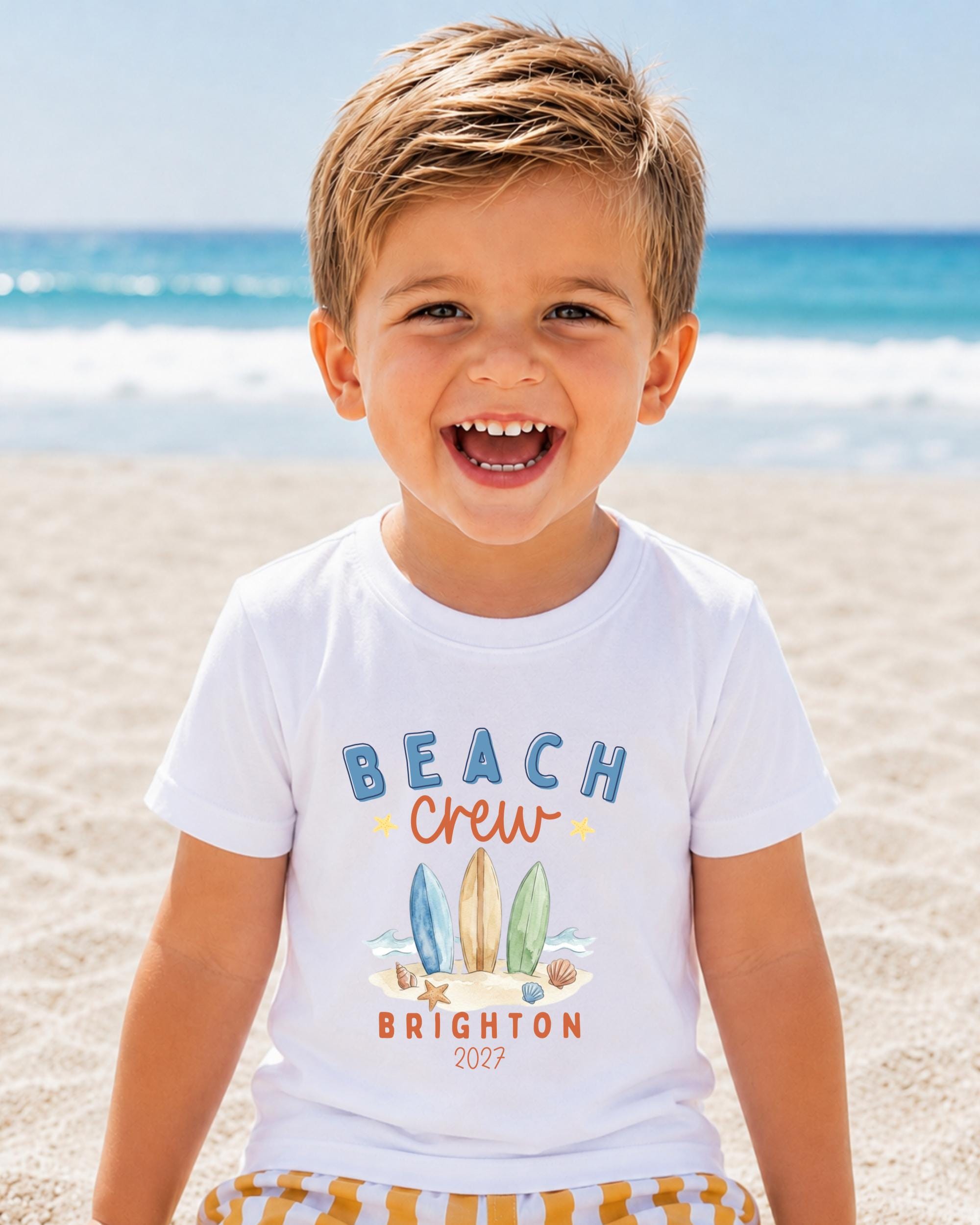 A young boy with a big smile on his face, wearing a white t-shirt with a beach crew design and text, standing on a sandy beach with the ocean in the background.