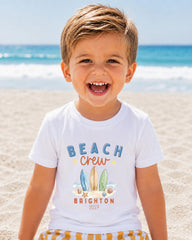 A young boy with a big smile on his face, wearing a white t-shirt with a beach crew design and text, standing on a sandy beach with the ocean in the background.