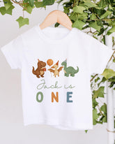 A white t-shirt with a graphic design featuring cartoon dinosaurs and the text "Jack is ONE" printed on it.