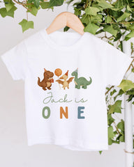 A white t-shirt with a graphic design featuring cartoon dinosaurs and the text "Jack is ONE" printed on it.