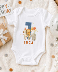 A white onesie with a personalized number 7 and animal designs, including a zebra, lion, and elephant, is displayed on a bed with a "Happy Birthday" sign and a balloon.