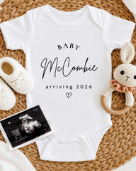 A white baby onesie with the text "BABY McCOMBIE arriving 2026" printed on it, along with a pair of white crocheted shoes and a stuffed rabbit toy.