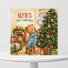 The image is a book cover for a children's book titled "Alfie's First Christmas". The cover features a teddy bear decorating a Christmas tree, with a cozy fireplace and other holiday decorations in the background.