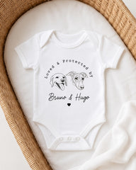 A white baby onesie with a graphic design of two dog faces and the text "Loved & protected by" above them, placed on a woven basket.