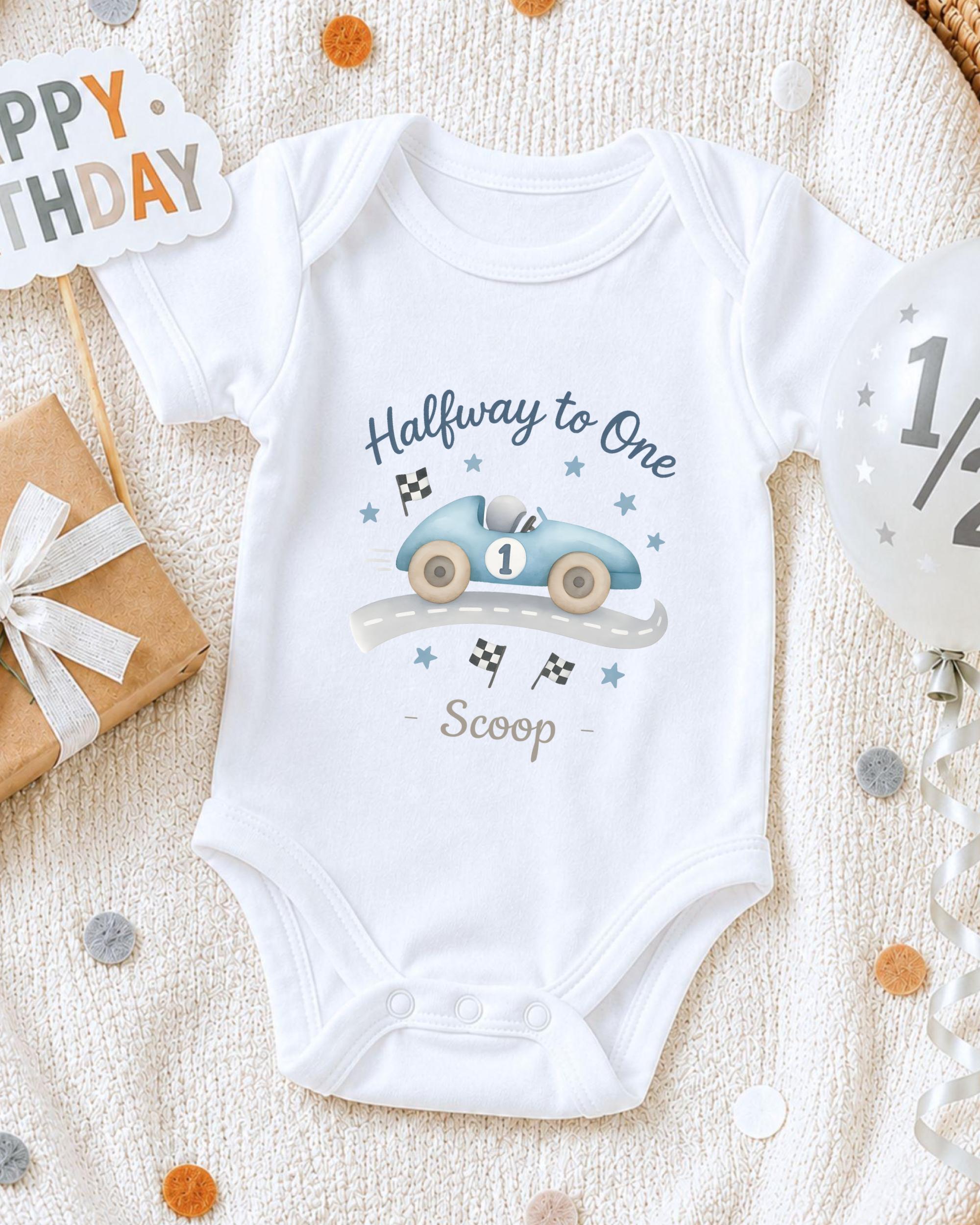 A white onesie with a race car design and the text "Halfway to One Scoop" printed on it, along with a "Happy Birthday" sign and a balloon.