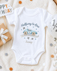 A white onesie with a race car design and the text "Halfway to One Scoop" printed on it, along with a "Happy Birthday" sign and a balloon.