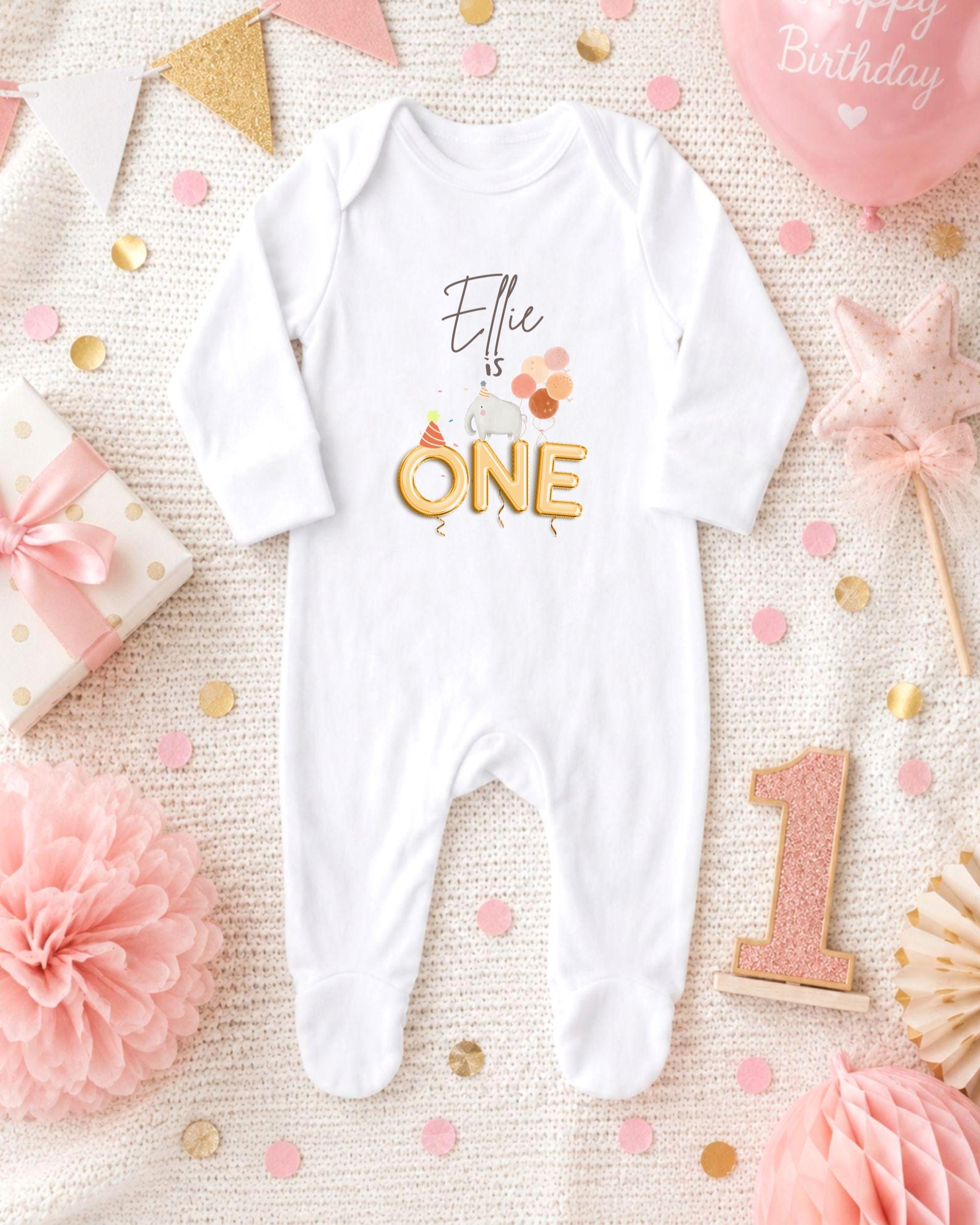 A white onesie with the text "Elle is one" printed on it, surrounded by birthday decorations such as a pink star, confetti, and a pink balloon.