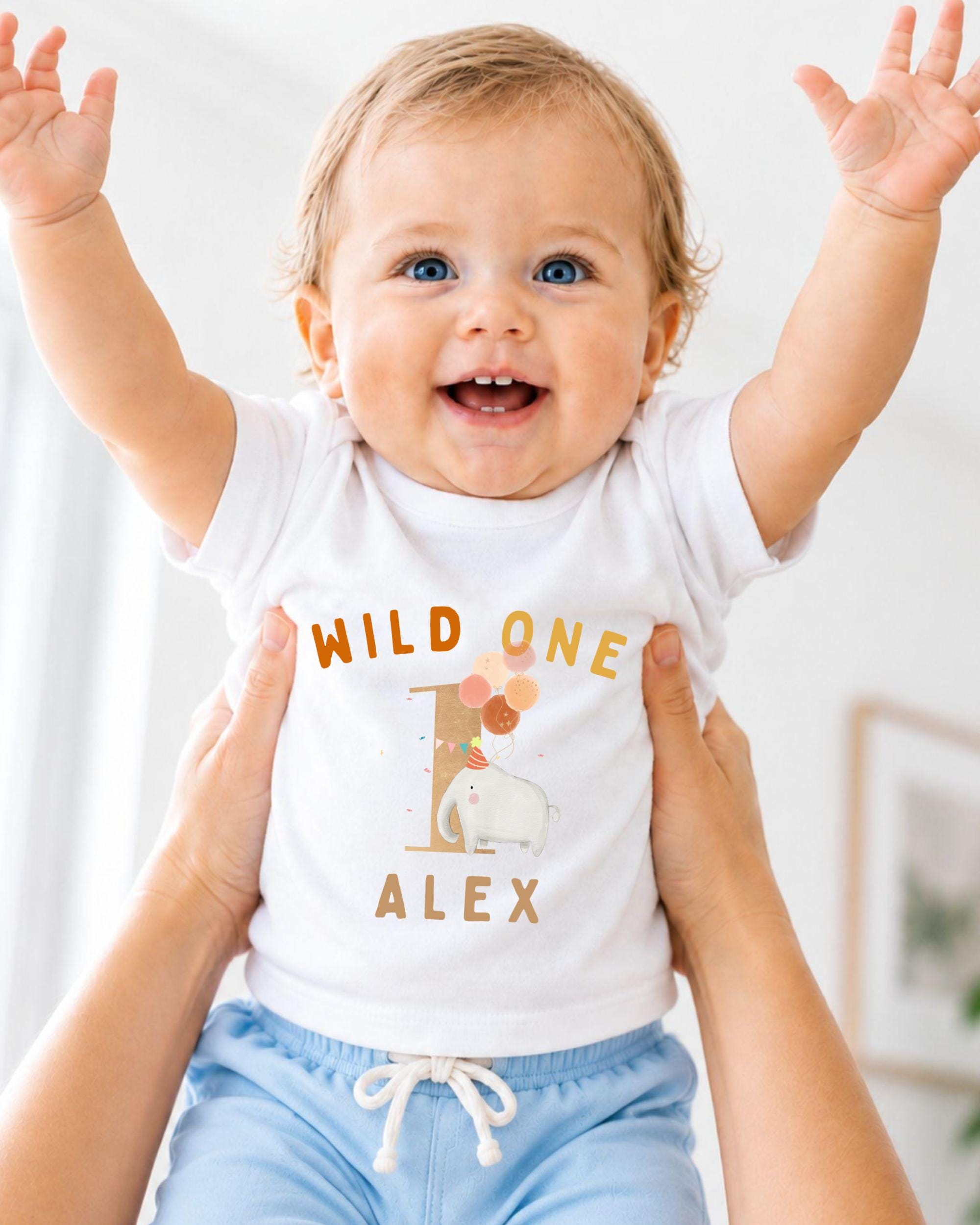 A smiling baby wearing a white t-shirt with the text "WILD ONE ALEX" and an elephant graphic, being held up by someone's arms.