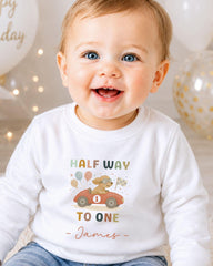 A smiling baby boy wearing a white shirt with a colorful graphic and text, sitting in front of birthday balloons.