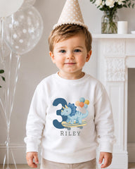 A young boy wearing a white sweatshirt with a dinosaur and balloons on it, standing in front of a fireplace with balloons and a vase of flowers.