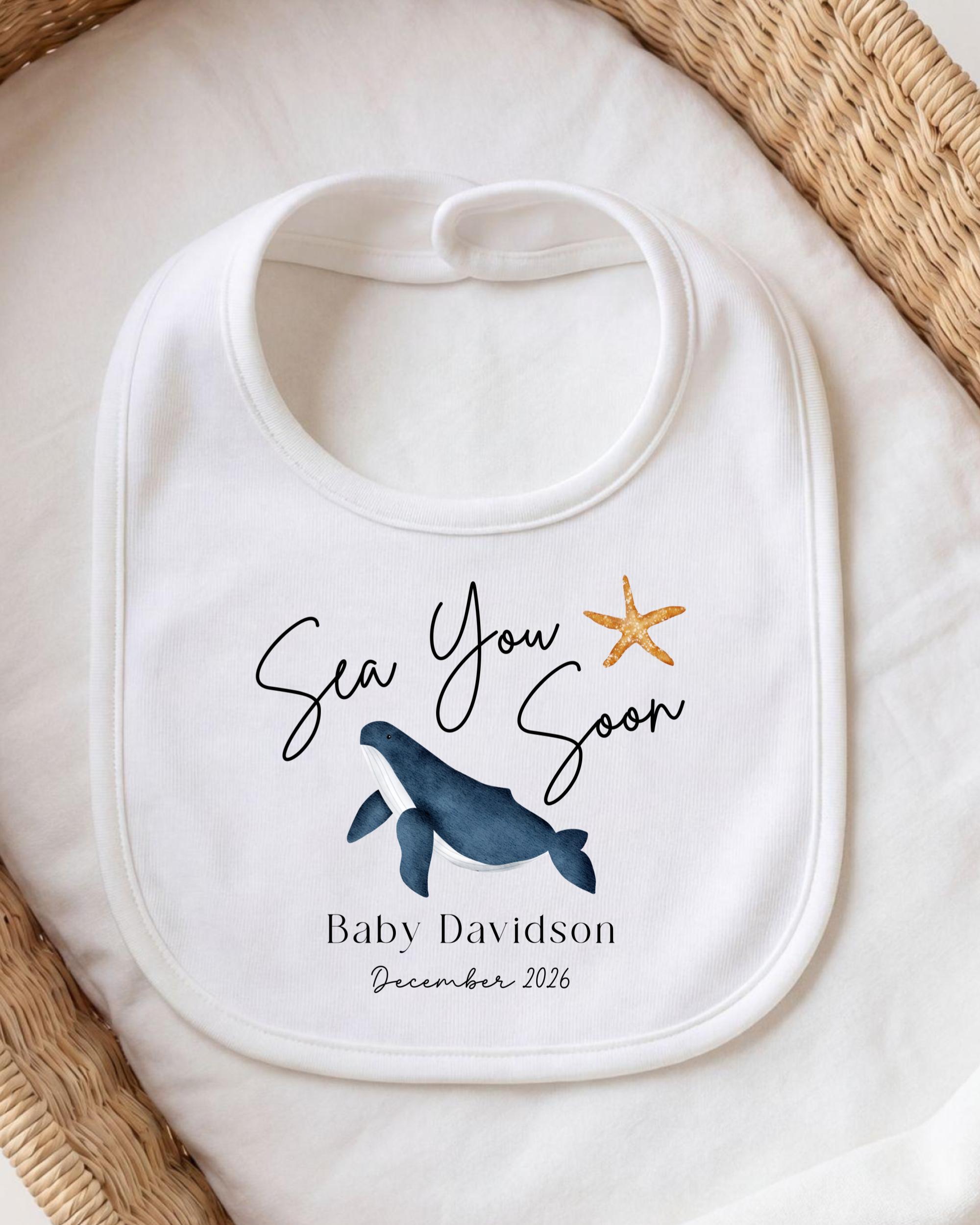 A white bib with a graphic of a whale and a starfish, along with the text "Sea You Soon" and "Baby Davidson December 2026".