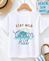 A white t-shirt with a graphic design of a wave and the text "STAY WILD Ocean kid" printed on it, hanging on a wooden hanger.