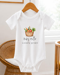 A white baby onesie with a basket of vegetables printed on it, hanging on a wooden hanger.