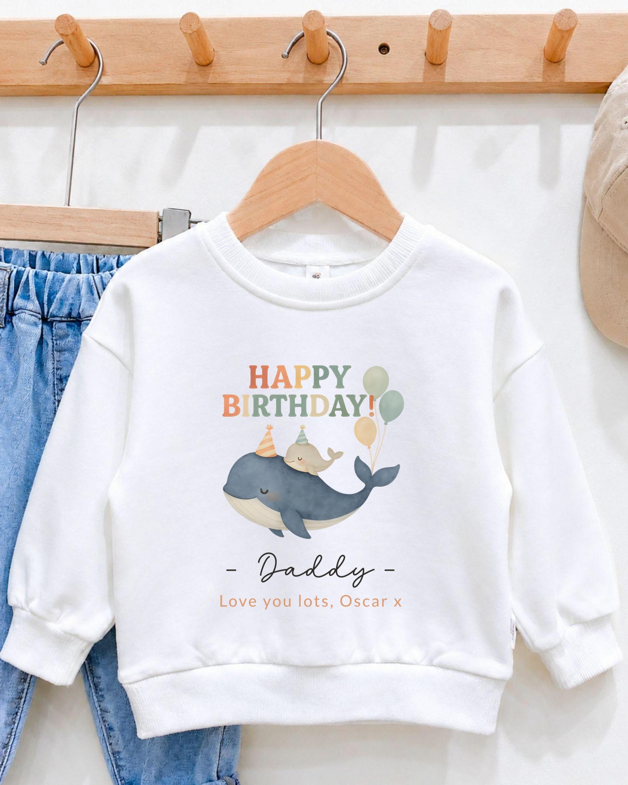 A white sweatshirt with a graphic of a whale and balloons, along with the text "Happy Birthday Daddy - Love you lots, Oscar x" printed on it.