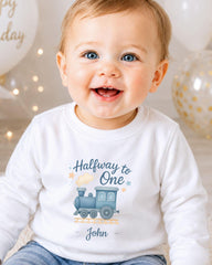 A smiling baby boy wearing a white shirt with a train graphic and the text "Halfway to One John" printed on it.