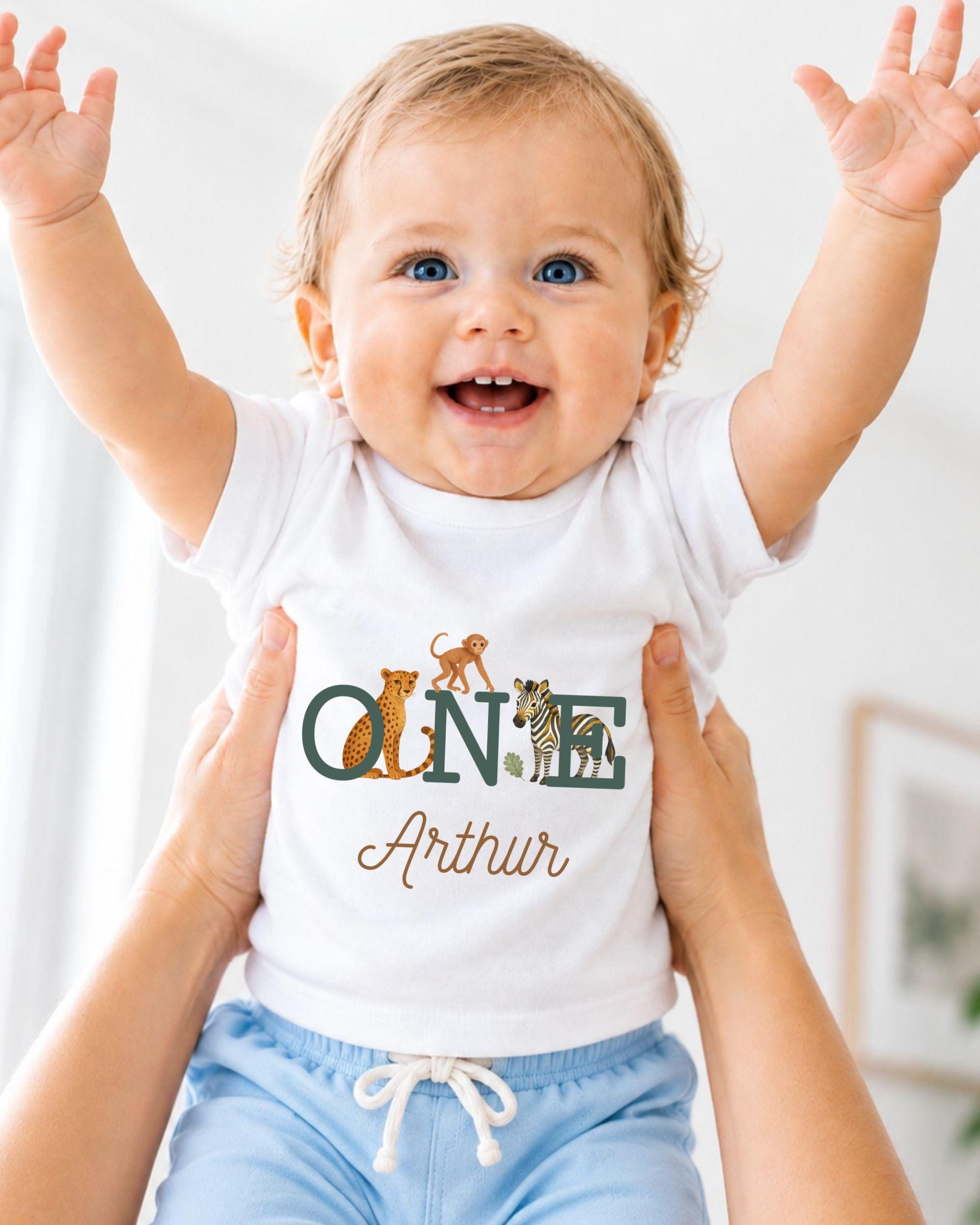 A smiling baby wearing a white t-shirt with the name "Arthur" and animal illustrations, being held up by someone's arms.