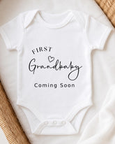 A white onesie with the text "FIRST Grandbaby Coming Soon" printed on it.