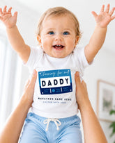 A smiling baby wearing a white shirt with a graphic design and text, being held up by someone's arms.