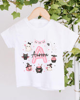 A white t-shirt with a personalized design featuring a pink "A" surrounded by various Halloween-themed illustrations, including a ghost, bats, a spider, a pumpkin, a witch's hat, a pumpkin, a cupcake, and a spider web.
