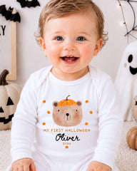 A smiling baby boy wearing a white onesie with a pumpkin and bear design, sitting in front of Halloween decorations.
