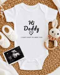 A baby onesie with the text "Hi Daddy" and a heart symbol, along with a crocheted bunny toy and a baby bootie, all arranged on a woven placemat.