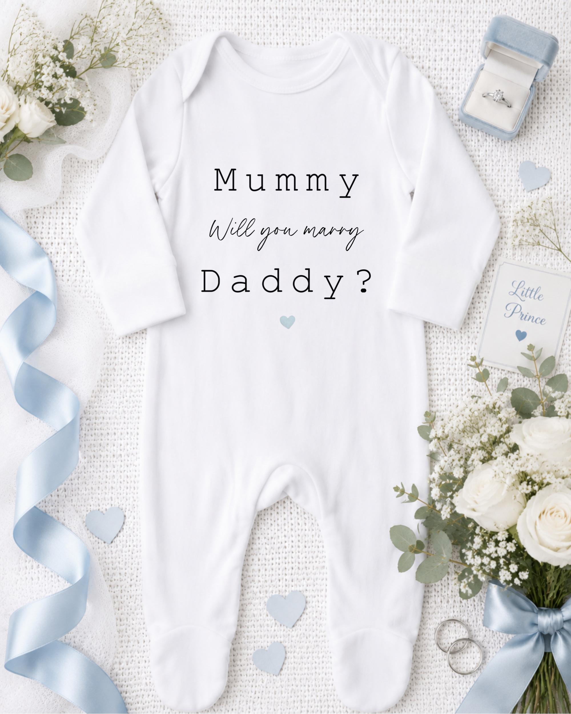 A white onesie with the text "Mummy, Will you marry Daddy?" printed on it, along with a ring, a bouquet of white flowers, and a blue ribbon.