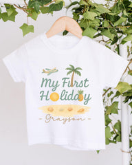 A white t-shirt with a graphic design featuring a palm tree, a plane, and various beach-related items, along with the text "My First Holiday - Grayson".